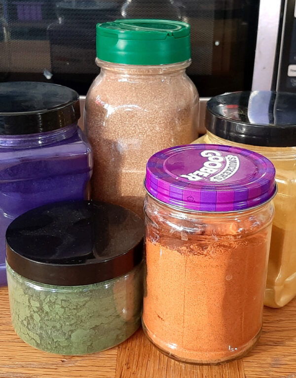 Five jars, some glass, some plastic, holding a variety of brightly colored powders.