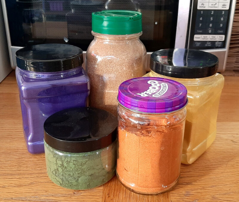 Five jars, some glass, some plastic, holding a variety of brightly colored powders.