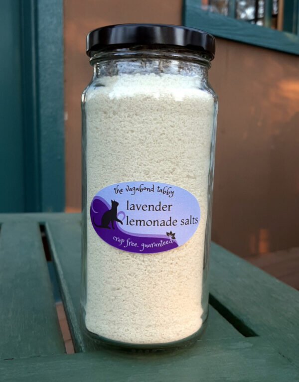 A tall, clear glass jar of bath salts, yellow with a layer of lavender flowers on top.