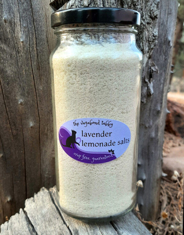 A tall, clear glass jar of bath salts, yellow with a layer of lavender flowers on top.