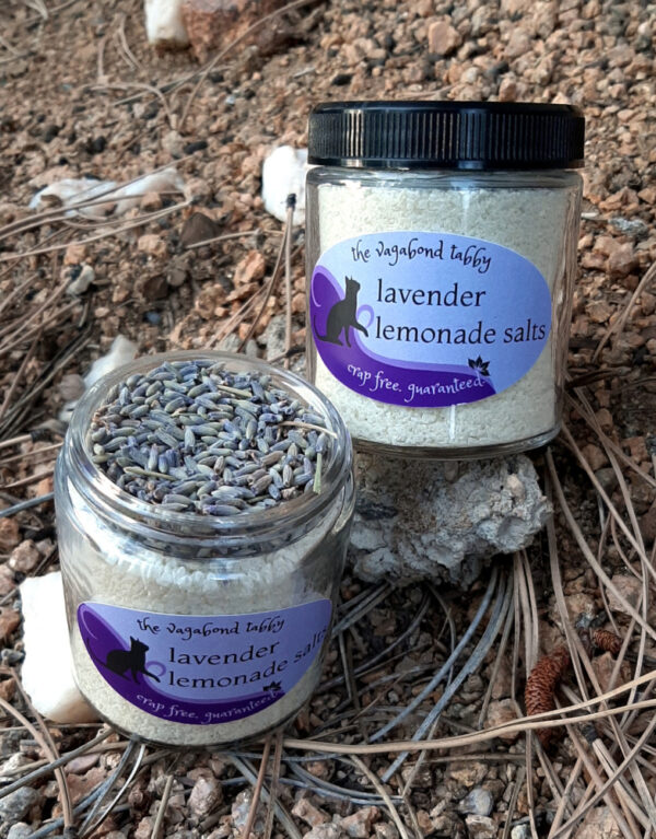 Two clear glass jars of bath salts, yellow with a layer of lavender flowers on top. One is open to show the lavender.