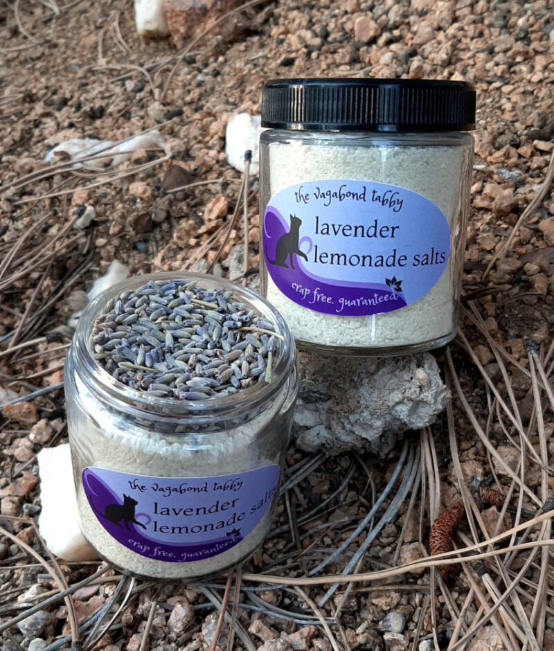 Two clear glass jars of bath salts, yellow with a layer of lavender flowers on top. One is open to show the lavender.