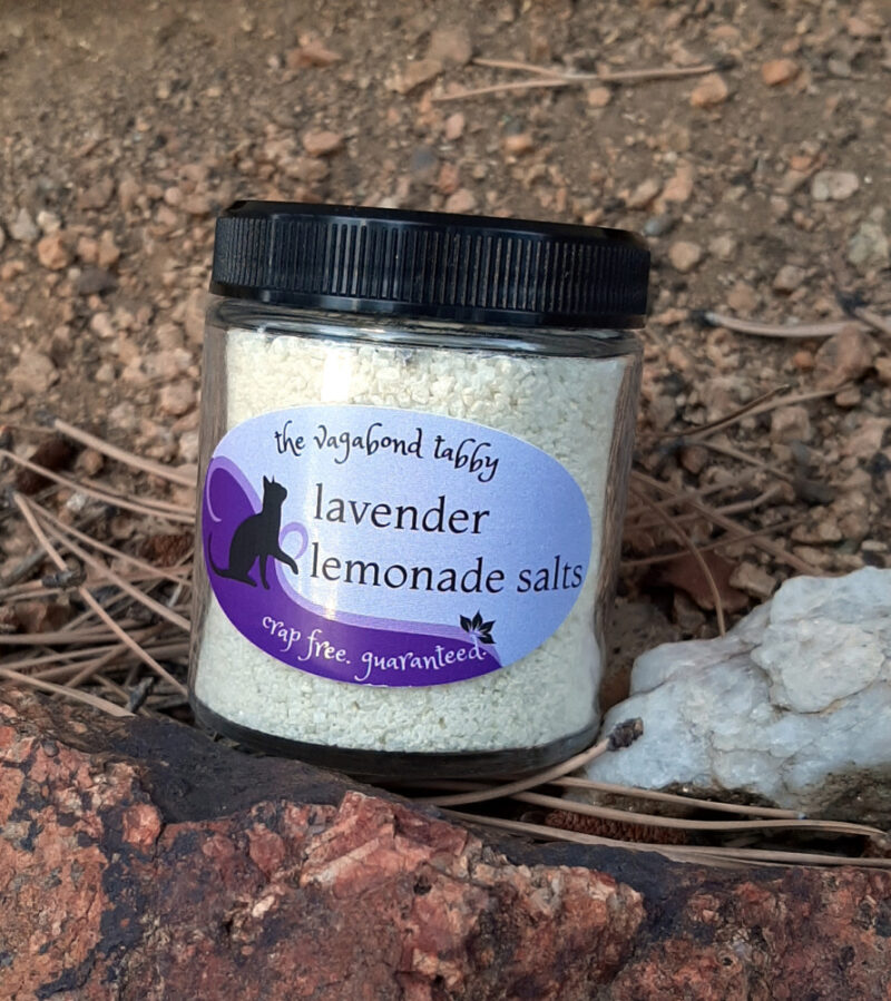 A clear glass jar of bath salts, yellow with a layer of lavender flowers on top.