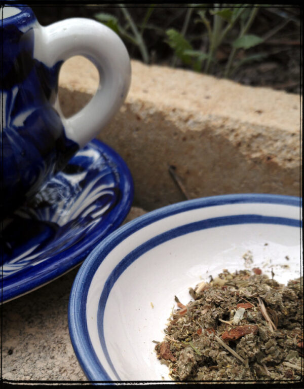 A blue and white pottery saucer holds a small heap of various herbs; next to it is a matching teapot, in its own saucer.