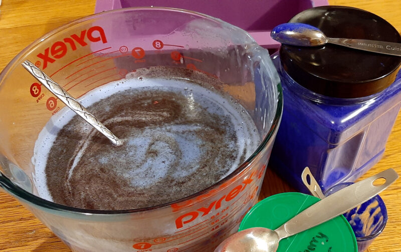 A large glass measuring bowl full of melted soap; a brown powder is being stirred into it and makes a nice swirl pattern.