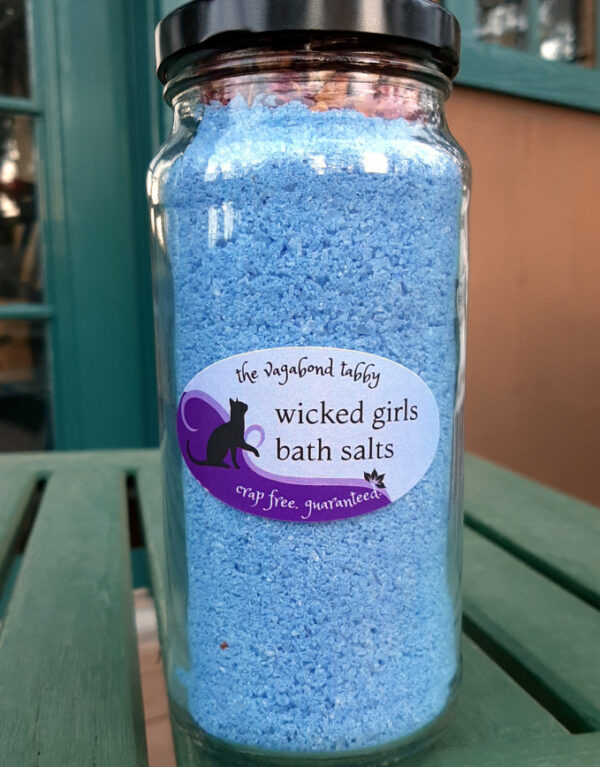 A tall clear glass jar filled with blue bath salts. The very top holds a layer of rose petals.