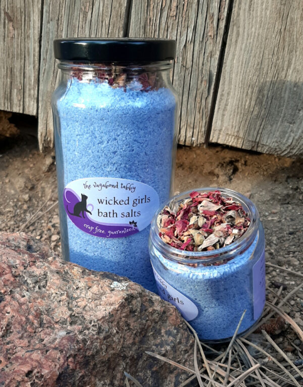 Two glass jars filled with blue bath salts. The shorter one is open, showing a layer of rose petals.