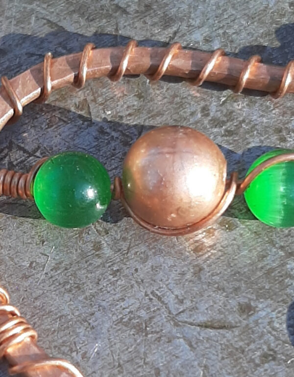 An asymmetrical arch in copper wire -- one end turned inwards, the other curved out. Hanging down the middle are two green glass beads with a copper one between them, all threaded onto thin copper wire.