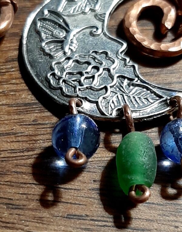 Hammered copper arches, one end spiraling inward while the other end curves out to a simple loop. Inside hang crescent moons, but broad, almost half moons, with butterflies and leaves embossed inside. From the bottom of each moon hang beads, blue green blue, each from its own loop.