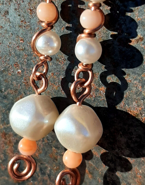 Each earring has two sections. The lower has a round white bead with a much smaller peach bead below it, both strung on copper wire with a neat spiral at the bottom. This is linked to a piece with another peach bead between two white beads, all strung on copper wire that curves back up around the beads, then twists around itself.