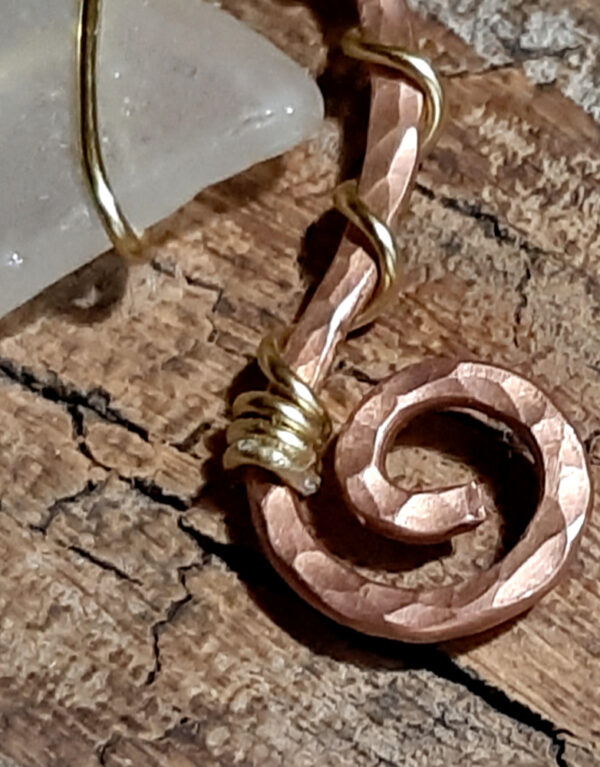 A piece of white creek glass is suspended between the arms of a rough arch of hammered copper wire, both wound round with much thinner golf-colored wire. One end of the arch ends in a spiral; the other extends further downward, and ends in a loop.