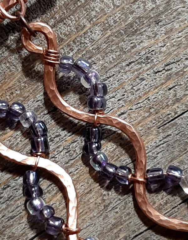 A broad horizontal bar of hammered copper wire, from which hang five meandering lengths of the same copper, each with purple and clear glass beads wired to them in complementary arcs.