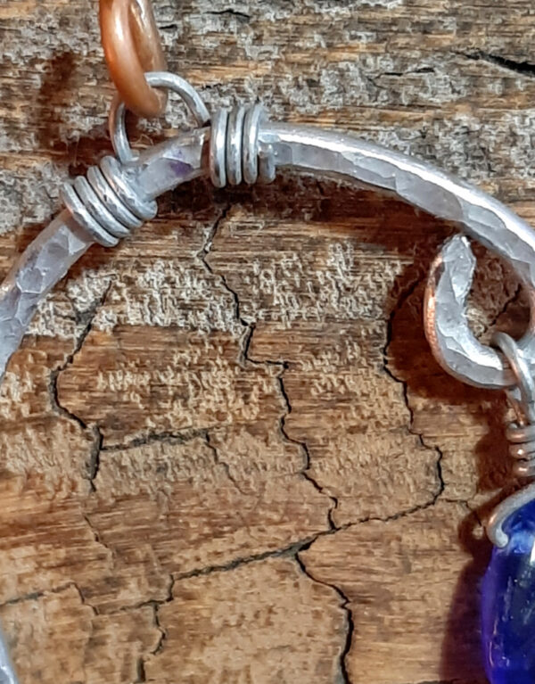 An uneven arc of hammered aluminum wire, with one end much higher than the other. From both ends hang cobalt blue glass leaves.