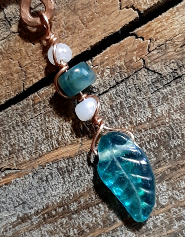 A squiggle of hammered copper wire with loops at top and bottom; from the bottom hangs a green glass leaf, strung on finer copper wire along with white and green beads.