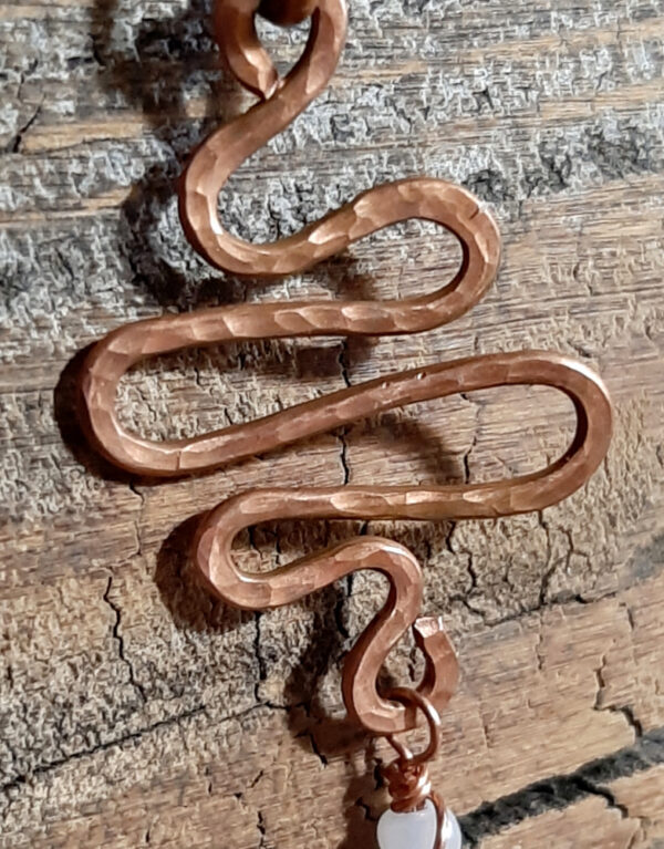 A squiggle of hammered copper wire with loops at top and bottom; from the bottom hangs a green glass leaf, strung on finer copper wire along with white and green beads.