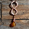 This pendant is a vertical infinity sign of hammered copper, with brown and amber-colored beads hanging below.