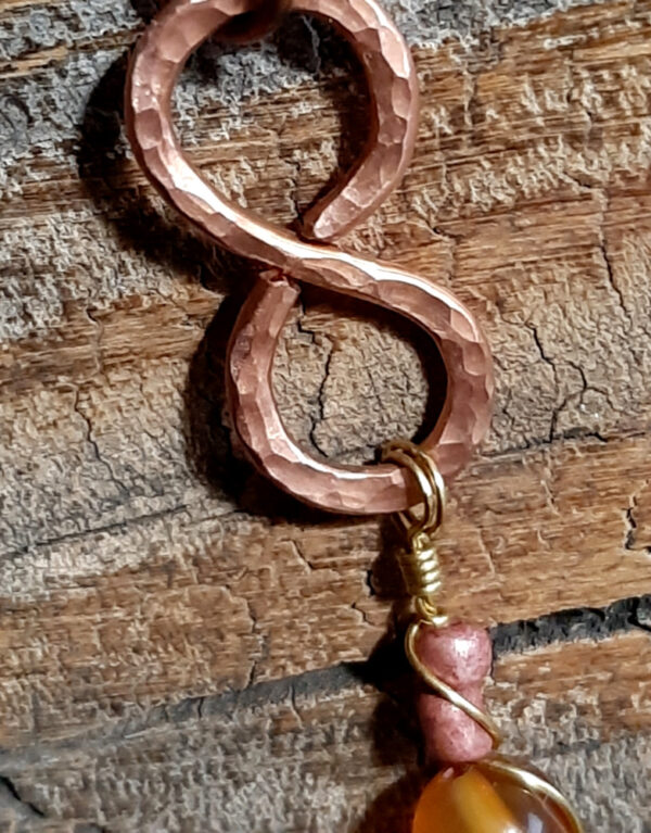 This pendant is a vertical infinity sign of hammered copper, with brown and amber-colored beads hanging below.