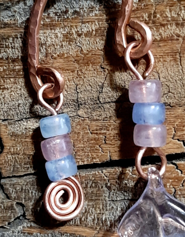 Each earring is an uneven arch of hammered copper, with three pink and pale blue beads hanging from the short end, and three pink and pale blue beads with a big clear, pink, and blue leaf hanging from the other.