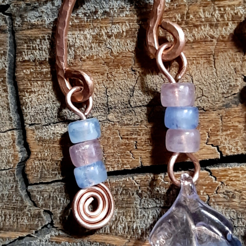 Each earring is an uneven arch of hammered copper, with three pink and pale blue beads hanging from the short end, and three pink and pale blue beads with a big clear, pink, and blue leaf hanging from the other.